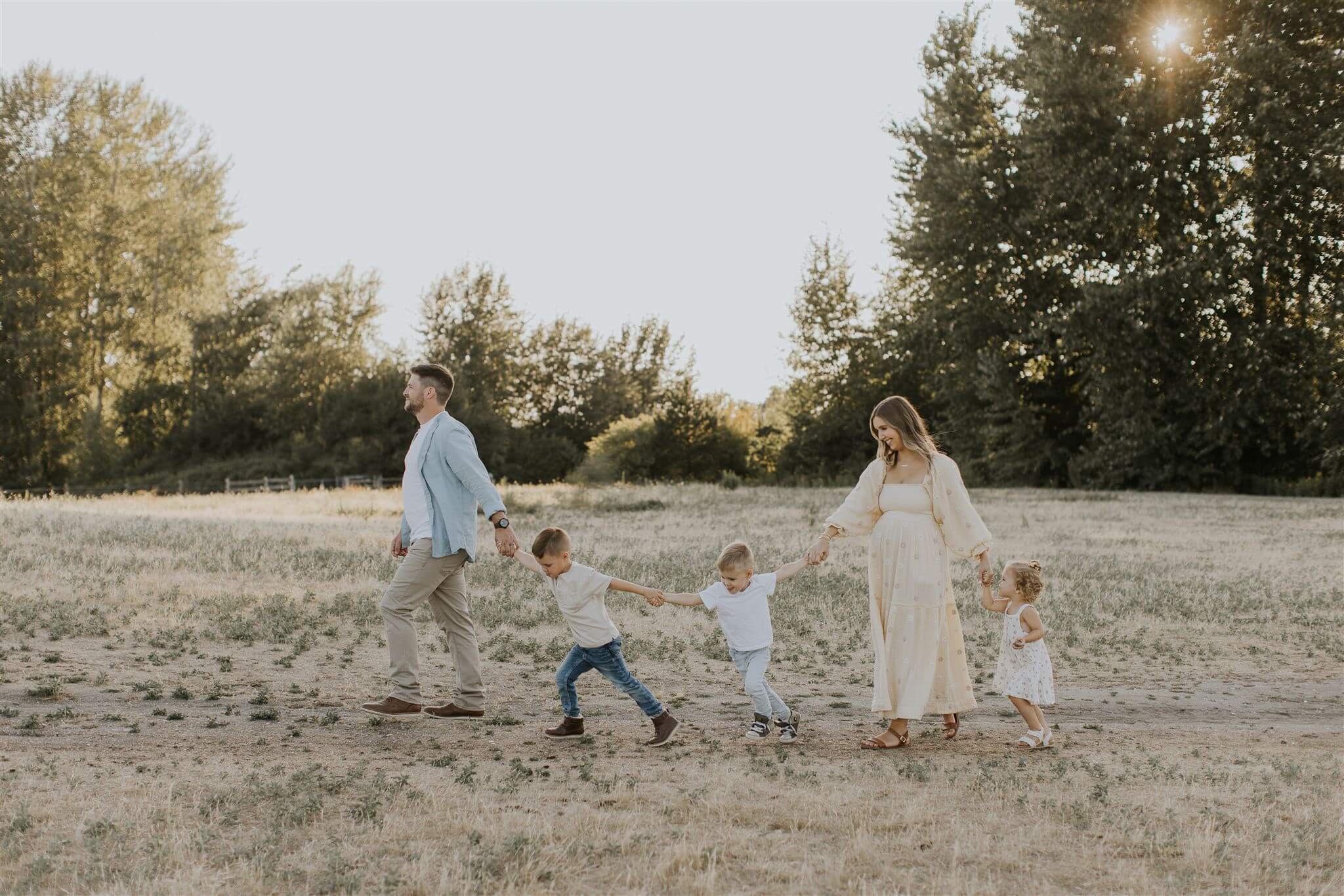 a young family of two boys and a girl with a pregnant mom walk in a line holding hands while the sun shines through the trees being them