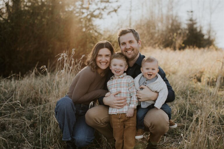 parents with two small boys crouch down in tall grass and smile at the camera