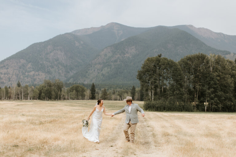 a bride and groom walk through a field and laughing at Bull River Guest Ranch in Cranbrook, BC