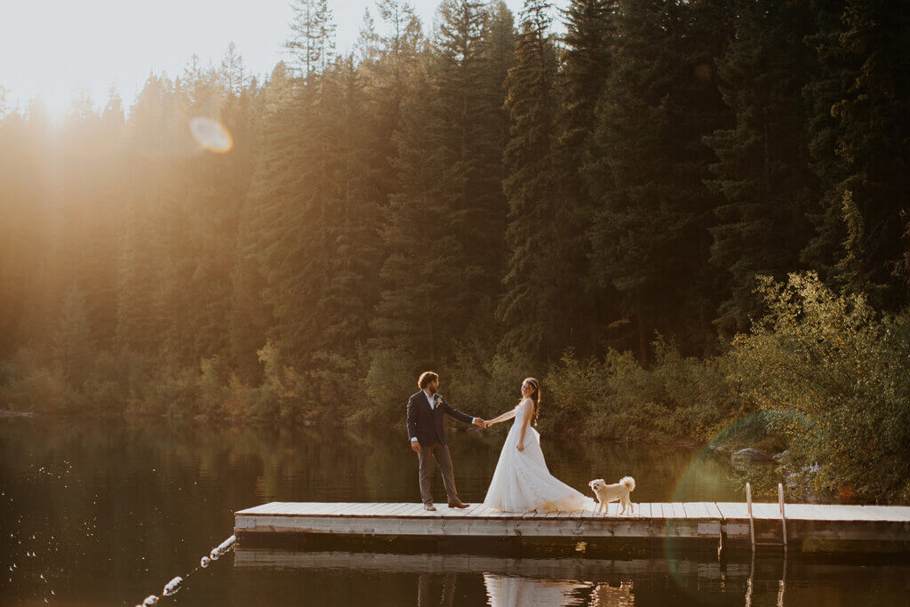 Bride and groom holding hands on a wooden dock by a lake, with a dog beside them and trees in the background.