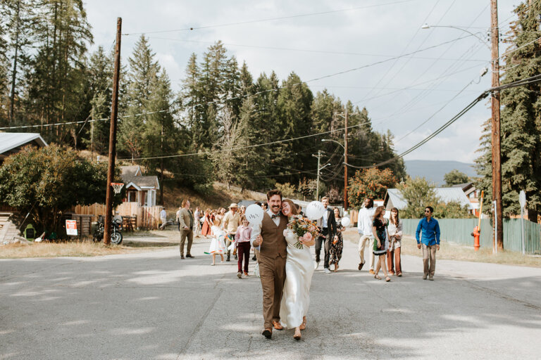 Couple walking together on a street, holding balloons and a bouquet, with guests in the background at a wedding celebration.