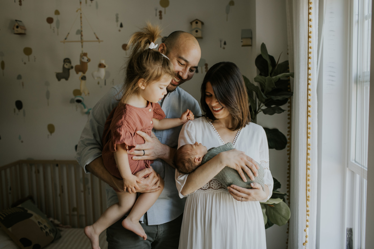 In-home newborn lifestyle scene in Kimberley, BC: a smiling mother holding a newborn, with a father and an older child nearby, soft natural light.