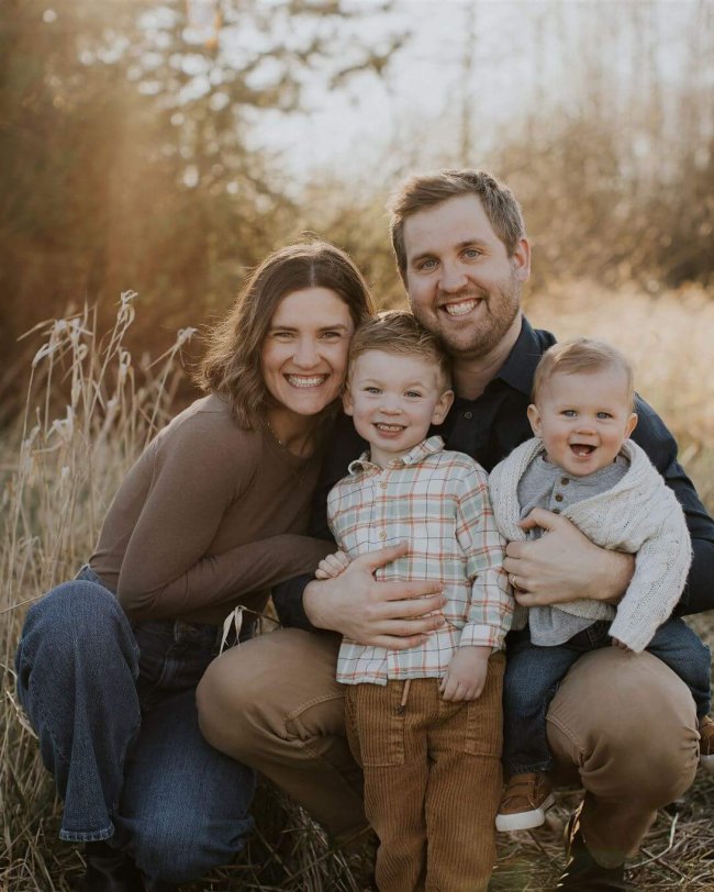 parents with two small boys crouch down in tall grass and smile at the camera