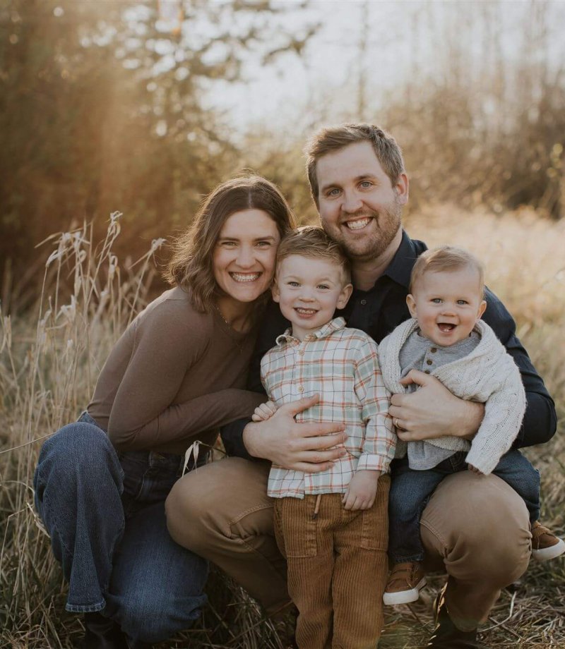 parents with two small boys crouch down in tall grass and smile at the camera