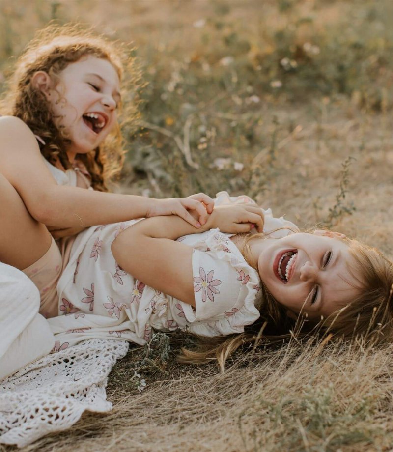 young girl laughing and tickling her smiling little sister on the ground in the warm glow of a sunny field