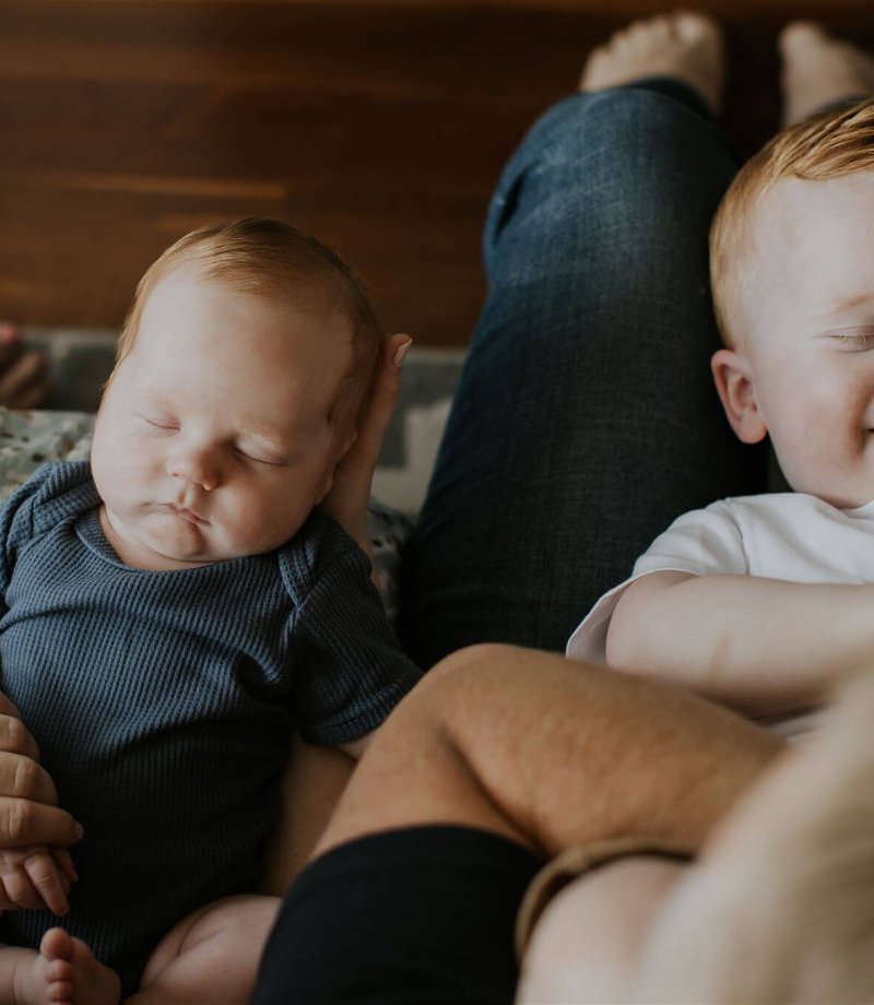 A little boy laughs on his dad's lap next to his newborn brother