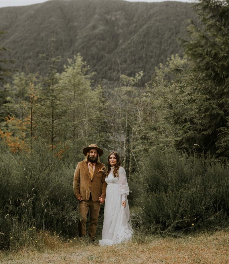 A couple in seventies inspired wedding attire look at the camera while standing amongst tall green grass in front of tree filed mountain in Cowichan Lake, British Columbia