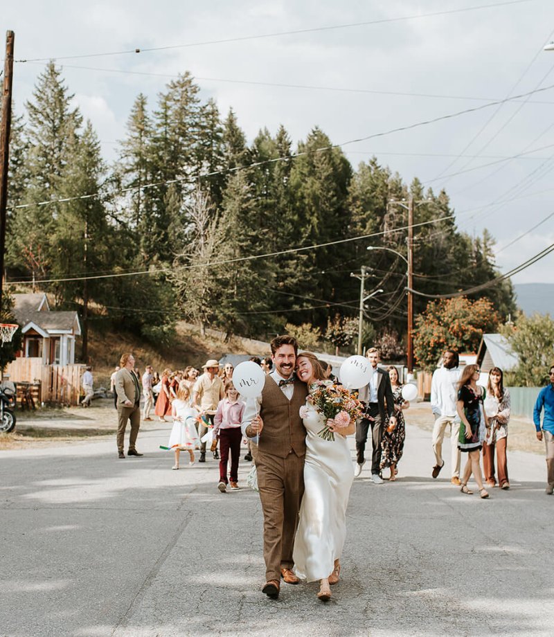 Couple walking together on a street, holding balloons and a bouquet, with guests in the background at a wedding celebration.