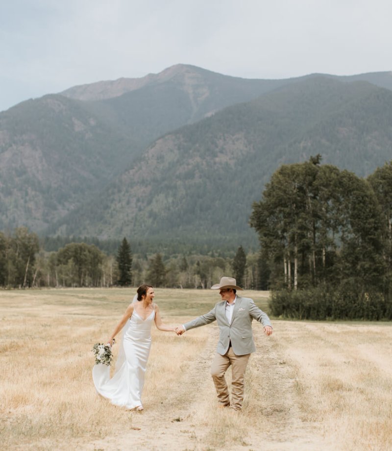 a bride and groom walk through a field and laughing at Bull River Guest Ranch in Cranbrook, BC